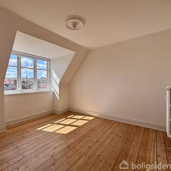 Et tomt værelse med parketgulv. Sollys strømmer ind gennem et stort vindue med udsigt til blå himmel og skyer, der kaster skygger på gulvet. En radiator står ved væggen.