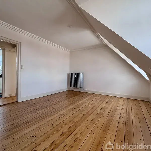 Et tomt værelse med skråvægge, trægulv og en radiator på væggen. Døråbning fører til et rum med en vaskemaskine, synligt i baggrunden.