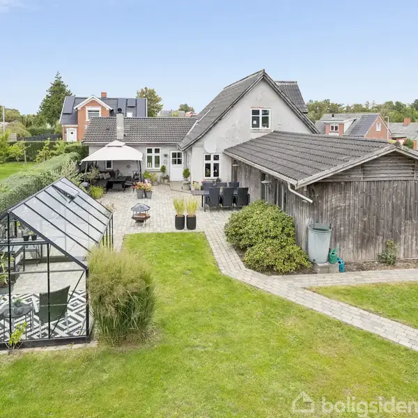 En stor, hvid villa med gråtag ligger forenden af en stenbelagt gårdsplads. Til venstre er et drivhus. Haven er grøn og omgivet af buske og træer.