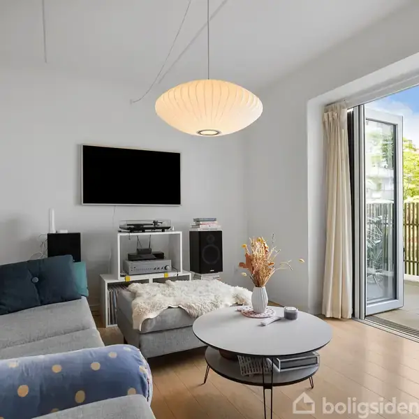 En lys stue med grå sofa og runde borde. Et sort tv hænger på væggen, mens havedøren står åben til en altan med grøn udsigt.