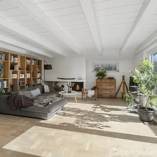 Stue med sofa og sofabord placeret ved siden af en stor træbogreol. Vinduerne lukker sollys ind, og planter dekorerer rummet. En kommode og stol står langs væggen.
