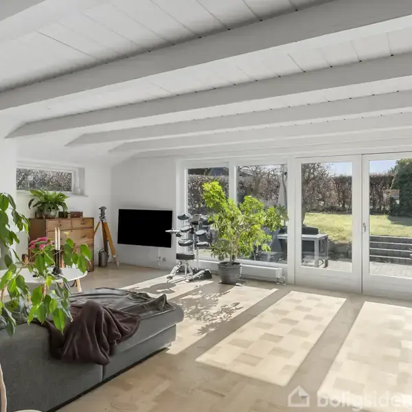 Stue med grå sofa og grønne planter, oplyst af naturligt lys fra store vinduer, der åbner op til en have. Hvidmalet loft med synlige bjælker og parketgulv.