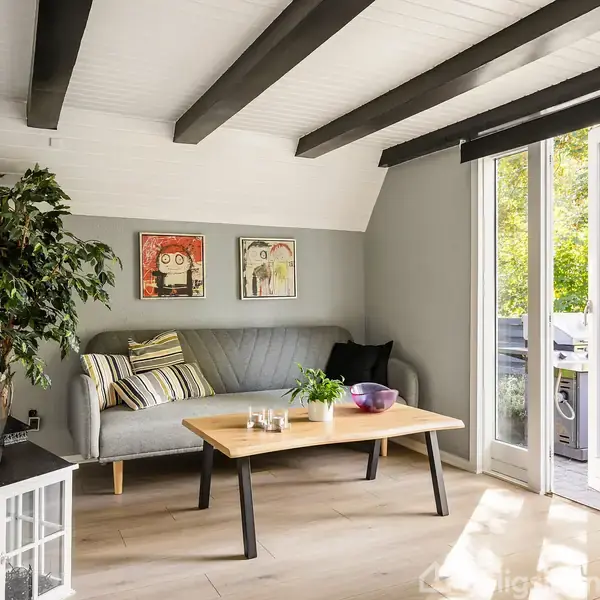Sofa står i en stue med lyse vægge, dekoreret med puder og billeder. Et sofabord står foran, og en stor plante ved siden af. En glasdør åbner til en terrasse med udsigt til træer.