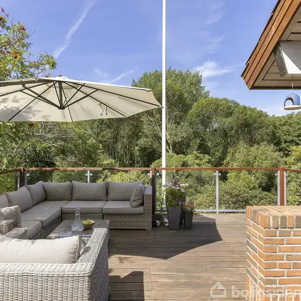 Grå hjørnesofa står på en træterrasse, dækket af en stor parasol. Terrasse omgivet af glasrækværk med udsigt til frodig have og blå himmel.