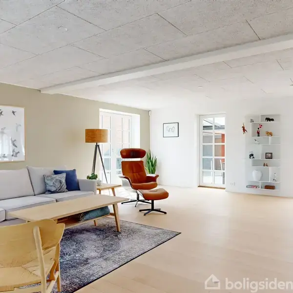 Sofa og lænestol i moderne stue, omgivet af lyse farver og dekorationer, med et stort vindue, der lader lys strømme ind. Kunst på væggen og et rundt tæppe.