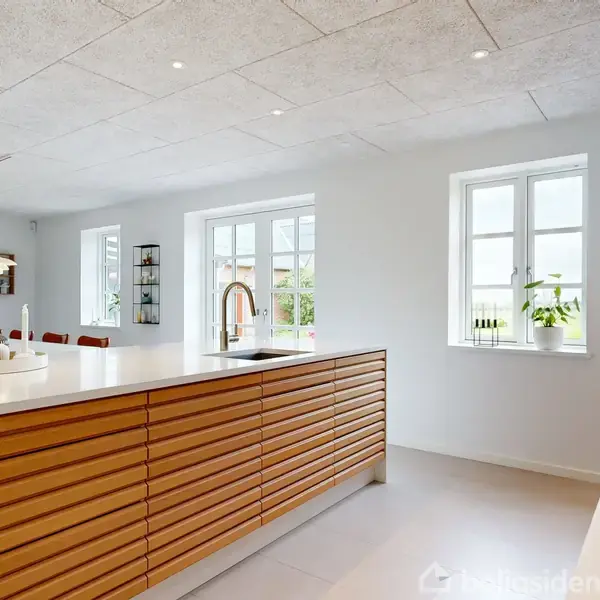 Et moderne køkken med en central ø, der har en trælamel forside. En vask og frugtskål står ovenpå. Store vinduer lader naturligt lys strømme ind i det åbne rum.