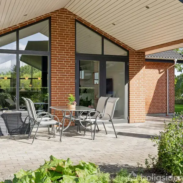 Et sæt havemøbler står på en terrasse foran et hus med røde mursten og store vinduer. Omkring er der grønne planter og en lille rød bygning i baggrunden.