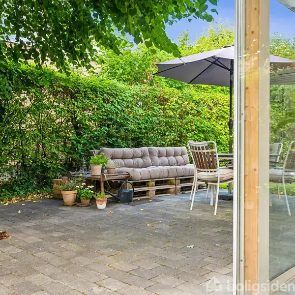 En sofa lavet af træpaller med puder står på en terrasse. Omkring står flere potteplanter, og en parasol giver skygge. Terrassen er omgivet af en grøn hække.