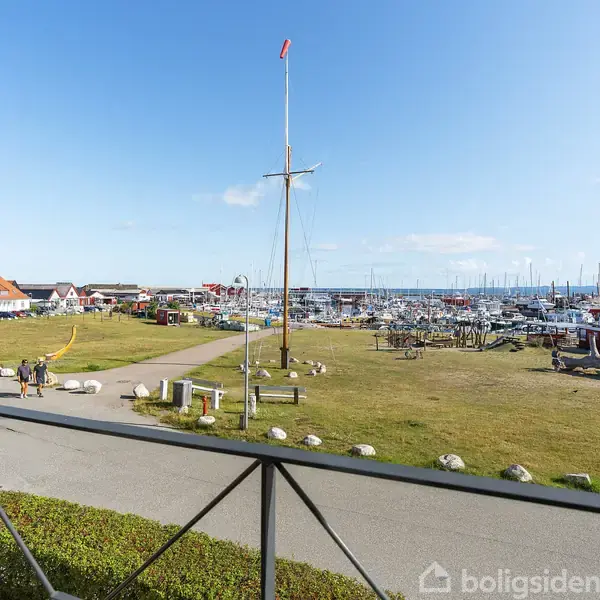 Udsigt over en havn med både og mast, hvor de røde tage af huse omkranser området. En sti går langs græsplænen, og mennesker går afslappet i solskinsvejr.