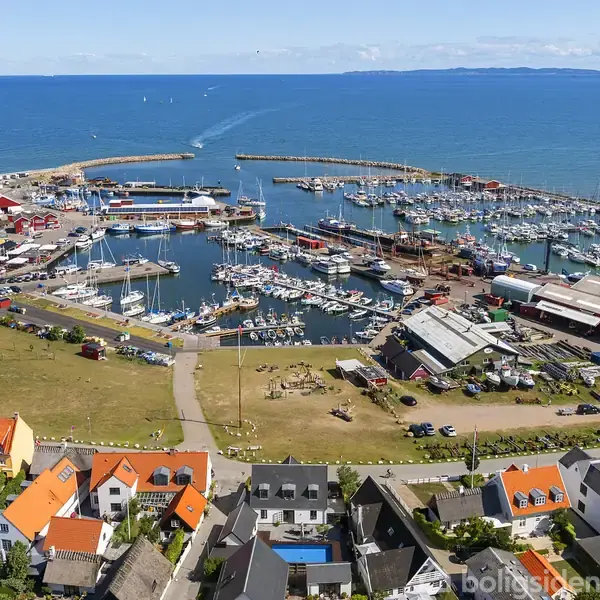 Havneområde fyldt med både ligger for anker i en travl marina. Omkringliggende bygninger med farverige tagrygge skaber en livlig atmosfære ved kysten. Baggrunden viser havet og en blå horisont.