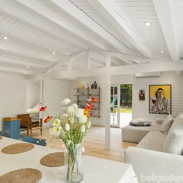 Stue med hvide trævægge og loftsbjælker. En stor, hvid sofa står ved en væg med maleri. Spisebord med blomster i forgrunden. Hylder og farverig reol fyldt med bøger.