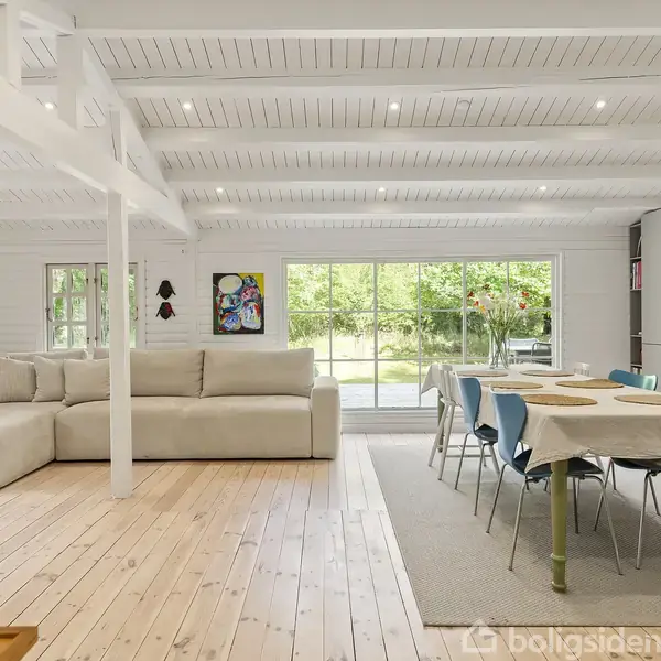 En lys stue med en L-formet sofa placeret ved siden af et spisebord med blå stole. Store vinduer giver udsigt til en grøn have. Vægge og loft er hvidmalede.
