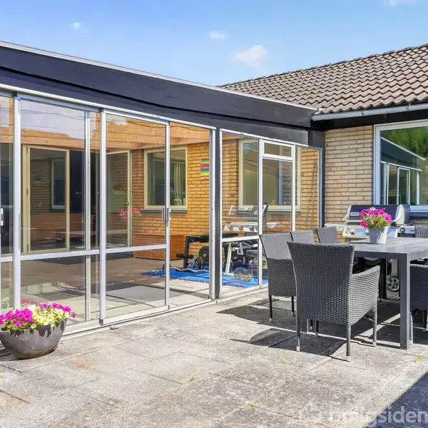 En overdækket terrasse med glasvægge indeholder et bord og stole. Ved siden af er et udendørs flisebelagt område med et spisebord og stole, omgivet af murstensvægge og et vindue.