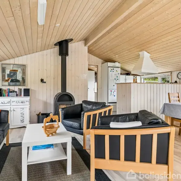 Sofaer og lænestol omkring et sofabord i en lys stue med brændeovn. Til højre står et spisebord med stole, mens et køkken ses i baggrunden. Trævægge og lofter omgiver rummet.