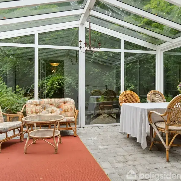 Veranda med glasvægge og glasloft. To kurvestole og et lille bord står til venstre på et rødt tæppe, mens et spisebord med fire stole er til højre. Lush have udenfor.