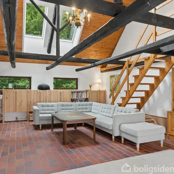 En moderne stue med en grå hjørnesofa står på et mørkt flisegulv. Trappen fører op til en hems under loft til kip med træbjælker. Pejs og reol står ved væggen.
