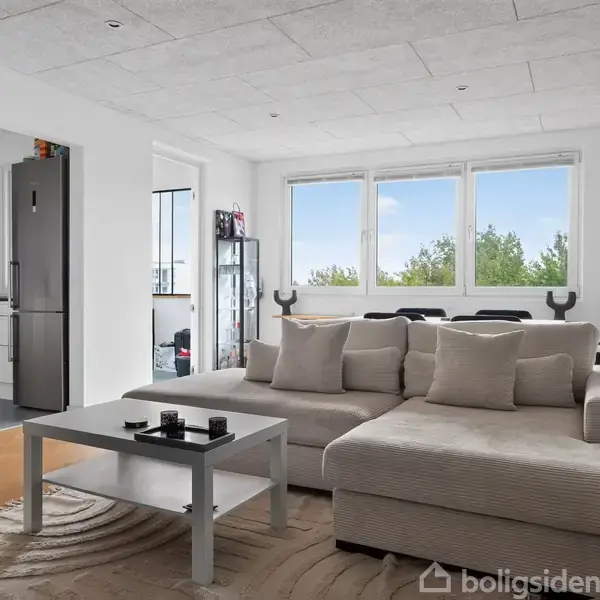 En lys stue med en stor beige sofa, der står foran store vinduer med udsigt til træer. Et minimalistisk sofabord står på et lysebrunt tæppe. En døråbning fører til et køkken med køleskab.