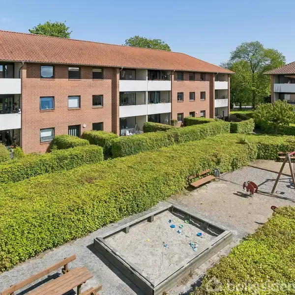 En legeplads med en sandkasse og gyngestativ er omgivet af velholdte grønne hække. Omkring ses murstensbebyggelser med røde tage under en klar blå himmel.
