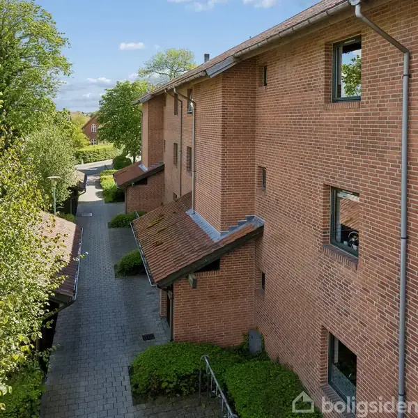 Bygning i røde mursten langs en flisebelagt sti, omgivet af grønne træer. Vinduerne er jævnt fordelt, og tagene har en let hældning. Vejret ser solrigt ud med blå himmel.