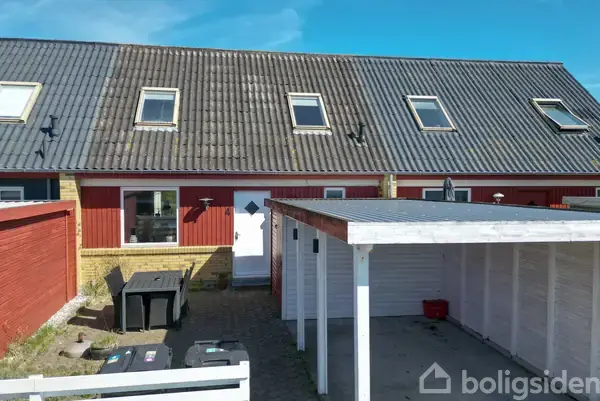 Rækkehus med røde træpaneler og skråt tag med fire ovenlysvinduer, står stille ved et hvidt skur i en gårdhave med brostensbelægning, på en solrig dag.