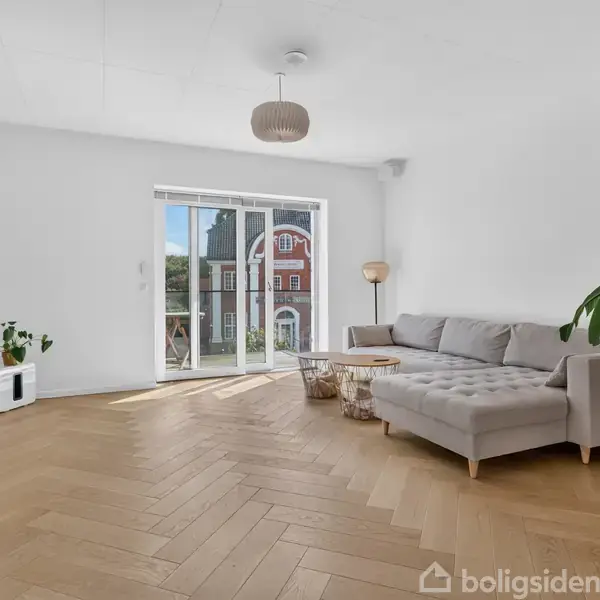 Stue med grå sofa, stort vægmonteret TV og sofabord. Store vinduer i baggrunden viser udsigt til en bygning. Grønne planter tilføjer liv til det minimalistiske rum.