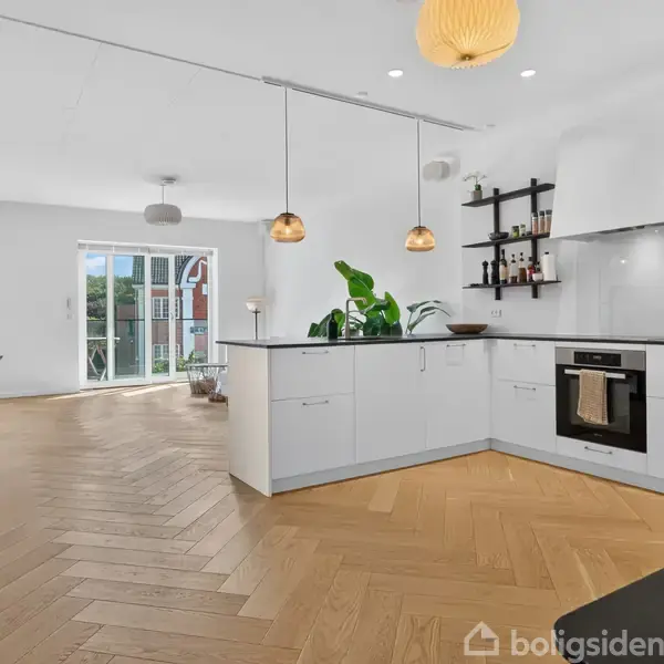 Et moderne køkken-alrum med hvide skabe, integreret ovn, og hængende lamper. Rummet har stort vindue, plante i hjørnet, og åbner ud til en altan med udsigt.