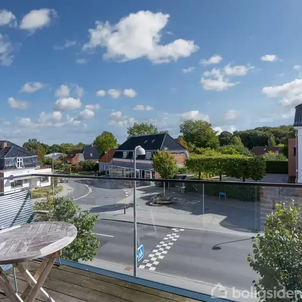 Et rundt bord og stol står på en balkon med glasrækværk. Udsigten viser en vej og bygninger i et solrigt byområde med blå himmel og skyer.