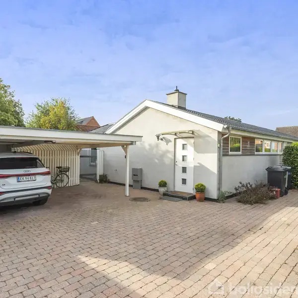 Hus med fladt tag står i et tilflugtsrum med carport. En hvid bil holder parkeret under carporten. Omkring huset er der brosten og grønne hække.