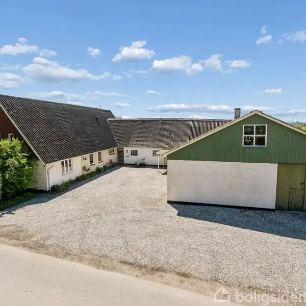 Et gårdkompleks med to bygninger, hvoraf den ene har grøn facade og garageporte. Bygningerne omkranser en gårdsplads med grusbelægning. Omgivelserne viser åbent landskab under en klar blå himmel.