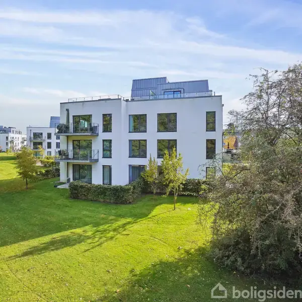 Hvid, moderne bygning står stille omgivet af grønne træer og en græsplæne i et solrigt bymiljø.