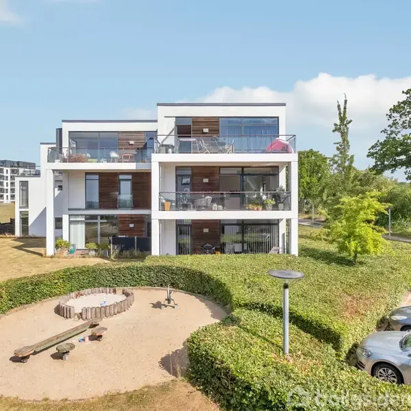 En moderne hvid lejlighedsbygning står i et grønt område. Der er terrasser med glasrækværk. Foran bygningen er der en sandlegeplads omgivet af hække, og en parkeringsplads til højre.