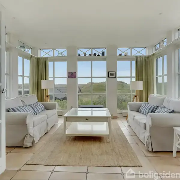 To hvide sofaer står overfor hinanden omkring et glasbord på et tæppe. Store vinduer omgiver rummet og slipper en masse naturligt lys ind, med landskab udenfor.