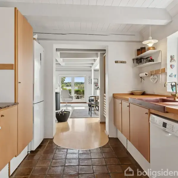 Køkken med træskabslåger og brun gulvflise, udstyret med komfur, køleskab og opvaskemaskine. Store vinduer til højre giver udsigt til en lys stue med møbler i baggrunden.