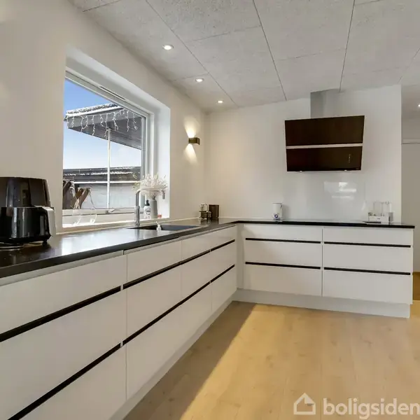 Et moderne køkken med hvide, grebsløse skabe og sorte bordplader. En køkkenmaskine står på bordet. Der er et stort vindue med udsigt til en terrasse. Væggene er hvide.