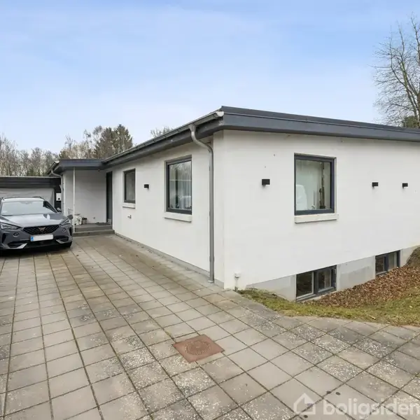 En moderne, hvid villa med fladt tag står på en flisebelagt indkørsel, hvor en bil er parkeret. Omkranset af hække og træer i et boligområde.