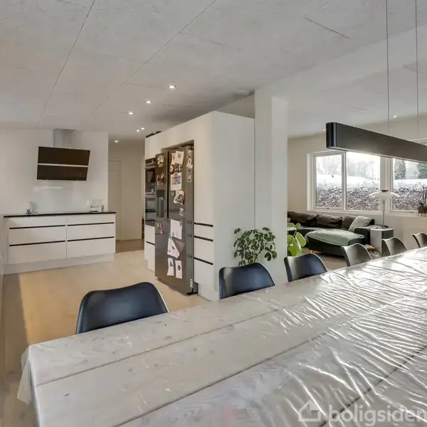 Spisebord dækket med dug står i forgrunden. Køkkenet med hvidt interiør ses bagved. En stor sofa og store vinduer i baggrunden. Blomsterdekoration på bordet.