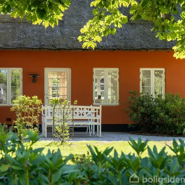 Et orange, stråtækt hus med hvide sprossede vinduer. En hvid bænk og bord står foran huset, omgivet af frodig have og grønne buske. Sollys skinner gennem træernes blade.