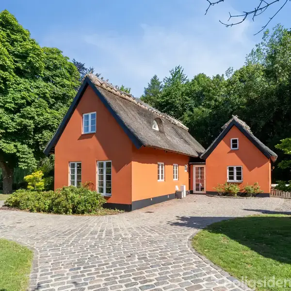 Et stråtækt, orange hus med hvide vinduer står midt i en brostensbelagt gårdsplads omgivet af grønne træer og buske. Vejret er klart og solrigt.