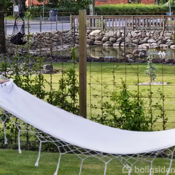 Hvid hængekøje hænger stille mellem to stolper i en have med grøn græsplæne og buske. I baggrunden ses et stengærde og en vej.