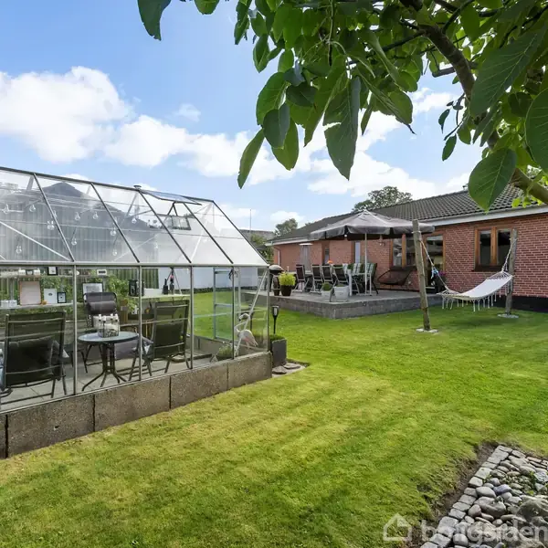 Drivhus står stille i en velholdt have ved siden af en rød murstensbygning. Plænen er frodig, og der er en terrasse med havemøbler og en hængekøje.