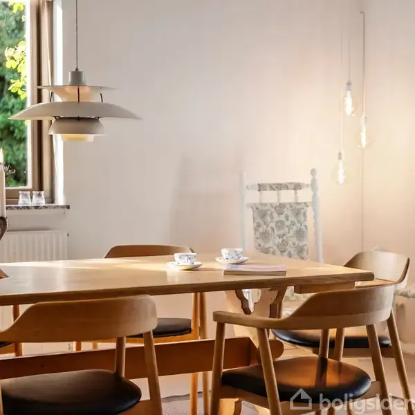 Et spisebord med træstole står i en spisestue. Bordet har stearinlys og kaffekopper. Rummet er oplyst af en moderne lampe og dekoreret med en plante ved siden af.