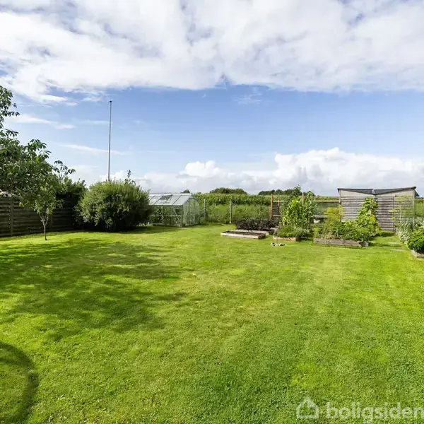 En stor, velplejet have med grøn græsplæne omgivet af enkelte træer og buske. Et drivhus og højbede er placeret langs et trådhegn under en blå, let skyet himmel.