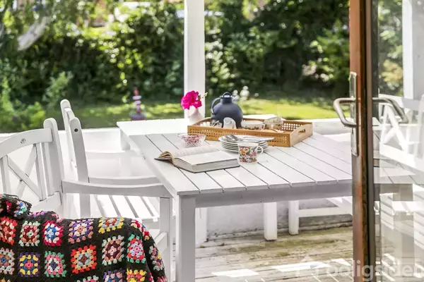 Et hvidt havebord med en farverig plaid ligger på en stol. På bordet står en bakke med en tekande, kopper, en bog og en blomst. Baggrunden er en grøn have.