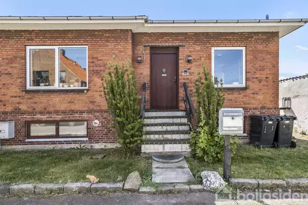 Murstenshus med trappe op til hoveddør omgivet af grønne buske. Til venstre og højre ses vinduer. I forgrunden en græsplæne, postkasse og affaldscontainere på asfalten.