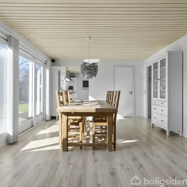Spisebord med seks træstole står i et lyst rum med store vinduer til venstre. En lampe hænger over bordet. Møbler inkluderer en hvid skænk. Trægulv og hvidmalede vægge.
