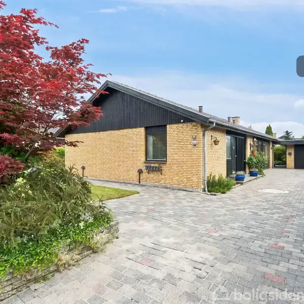 En gul murstensvilla med sorte trædetaljer ligger ved en flisebelagt indkørsel omkranset af en velplejet have med rødbladede buske og grønne planter. Tekst: “Visualisering”.