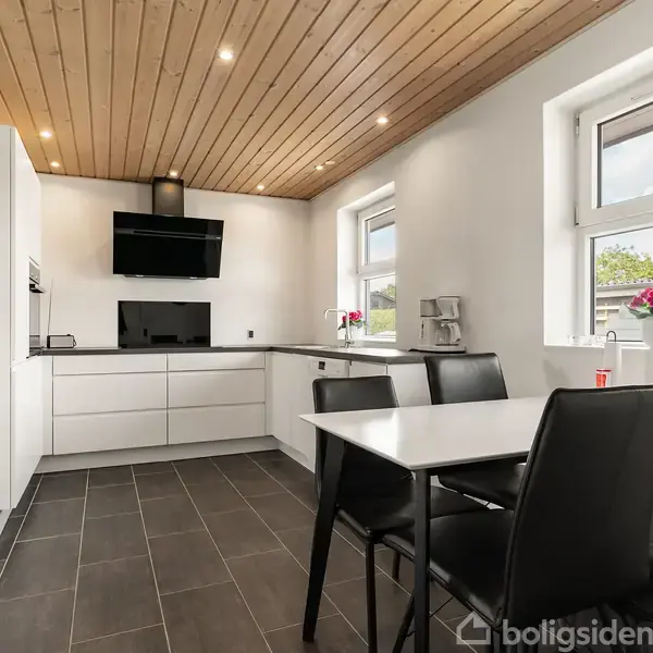 Et moderne køkken med hvide skabe og sort bordplade. Et sort spisebord med fire stole står foran et vindue. Træloft og flisegulv giver en elegant indretning.