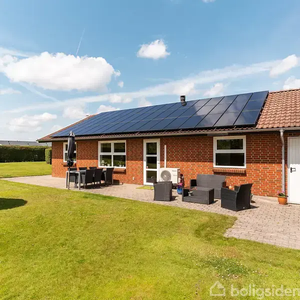 Rødt murstenshus med sort solcellepanel på taget, havemøbler på terrasse. Omgivet af grøn græsplæne og busk, klart himmel og få skyer i baggrunden.