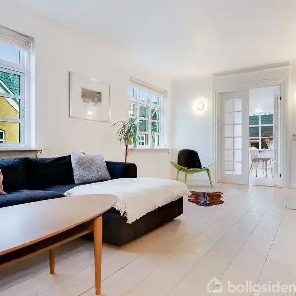 Sort sofa med puder står i en lys stue med trægulv, grøn stol og stort vindue. Dobbeltglasdør fører ind til et nærliggende rum. Grøn plante ved vinduet.