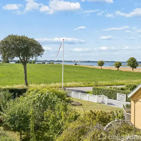 Et hus med gul facade står i forgrunden. Haven har et drivhus og frodig vegetation. I baggrunden ses grønne marker, en flagstang og en sø under en klar blå himmel.